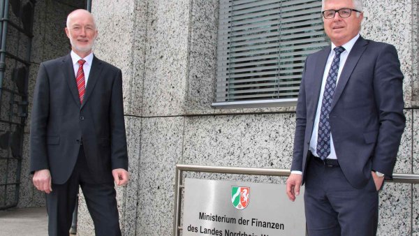 Zwei grauhaarige Männer in Anzügen stehen vor dem Schild des Finanzministeriums