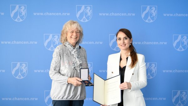 Zwei Frauen stehen vor einer blauen Wand und halten das Bundesverdienstkreuz und eine Urkunde 