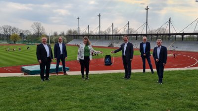 Staatssekretärin Andrea Milz steht mit 5 weiteren Menschen vor einer Sportstätte mit Tartanbahnen und einem Rasenplatz in der Mitte.