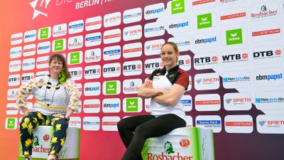 Staatssekretärin Andrea Milz sitzt mit einer Sportlerin vor einer großen Plakatwand.