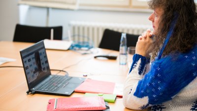 Staatssekretärin Milz im blau-weißen Pullover sitzt an einem beigen Besprechungstisch mit schwarzen Stühlen. Sie schaut vor sich auf einen schwarzen Notebook.