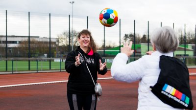 Frau Erika Rischko in weißer Daunenjacke und schwarzer Hose wirft der Staatsekretärin Andrea Milz in schwarzen Sportbekleidung einen bunten Sportland.NRW-Fußball zu. Die sechseckigen Felder des Fußballs haben die Sportland.NRW-Farben Weiß, Rot, Gelb, Grün, Blau und Dunkelblau. Auf dem Rücken tragt Frau Rischko einen schwarzen Sportland.NRW-Rucksack mit Sportland.NRW-Logo unten auf der Frontseite.