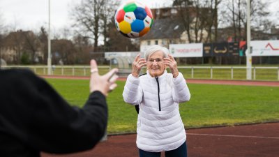 Frau Erika Rischko in weißer Daunenjacke und schwarzer Hose wirft der Staatsekretärin Andrea Milz in schwarzen Sportbekleidung einen bunten Sportland.NRW-Fußball zu. Die sechseckigen Felder des Fußballs haben die Sportland.NRW-Farben Weiß, Rot, Gelb, Grün, Blau und Dunkelblau.