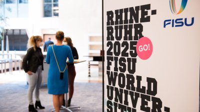 Drei Damen stehen draußen an einem runden Stehtisch. Rechts sieht man von der Rhine Ruhr 2025 FISU,.schwarz auf weiß.