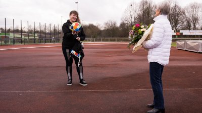 Frau Erika Rischko in weißer Daunenjacke und schwarzer Hose und Schuhen hält einen Blumenstrauß in ihren Händen. Ihr gegenüber stehend hält Staatsekretärin Andrea Milz in schwarzer Sportland-NRW-Sportbekleidung einen bunten Sportland.NRW-Fußball und einen Sportland.NRW-Rucksack in ihren Händen. Die sechseckigen Felder des Fußballs haben die Sportland.NRW-Farben Weiß, Rot, Gelb, Grün, Blau und Dunkelblau. Sie stehen auf einem Sportplatz.