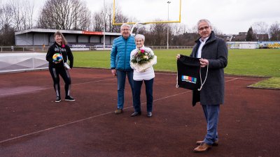 Frau Erika Rischko in weißer Daunenjacke und schwarzer Hose und Schuhen hält einen Blumenstrauß in ihren Händen. Neben ihr steht ein Herr in blauer Jacke. Links steh die Staatsekretärin Andrea Milz und ganz rechts der Bürgermeister von Langenfeld.