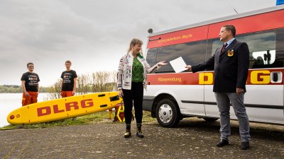 Eine Frau überreicht vor einem Rettungswagen eine Urkunde an einen Mann des DLRG