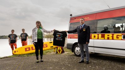 Ein Mann und eine Frau halten ein Tshirt hoch, im Hintergrund stehen zwei weitere Männer neben einem Rettungswagen