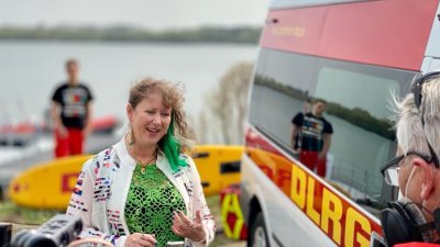 Eine Frau spricht vor einer Kamera, im Hintergrund ist ein Rettungswagen des DLRG