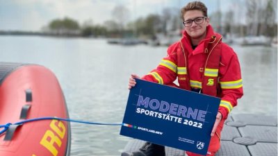 Ein Mann kniet auf einem Steg neben einem Schlauchboot und hält ein blaues Schild 