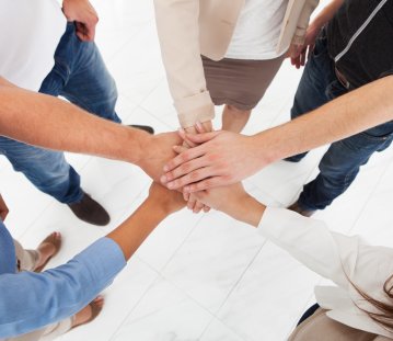 5 Menschen stehen im Kreis und haben jeweils ihre linke Hand in der Mitte sternförmig übereinander gelegt als Symbol für gute Teamarbeit.