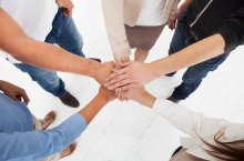 5 Menschen stehen im Kreis und haben jeweils ihre linke Hand in der Mitte sternförmig übereinander gelegt als Symbol für gute Teamarbeit.