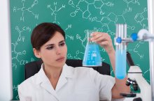 Eine dunkelhaarige Frau im weißen Kittel hält einen Glaskolben mit hellblauer Flüssigkeit in der linken Hand und schaut es prüfend an. Im Hintergrund eine grüne Tafel mit chemischen Formeln in weißer Kreideschrift.