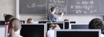 Auf dem Bild zu erkennen sind Schülerinnen und Schüler in einer Unterrichtsstunden am PC. Ein Lehrer schreibt Formeln an die Tafel.